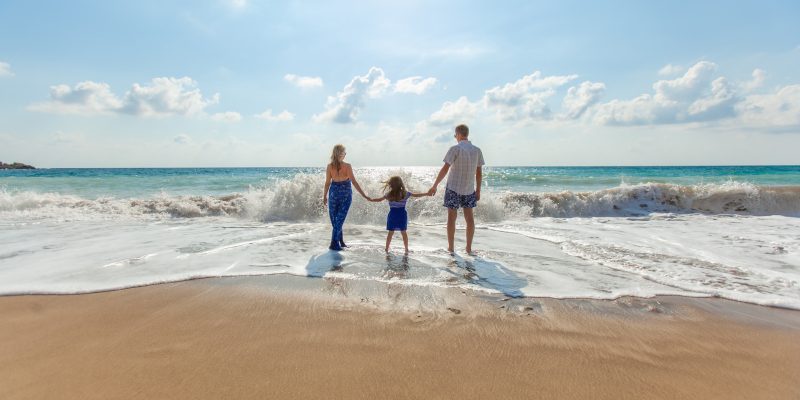 mother, child and father holding hands facing the beach.