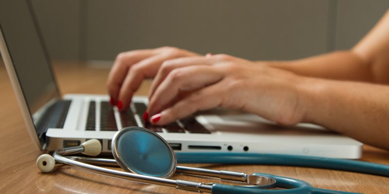 a laptop on a table while a hand is in the frame typing on the laptop while a stethoscope is in front of it.